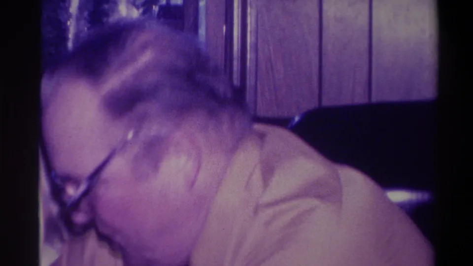 Close overhead shot of older man with heavy-framed glasses bent forward, motion blur from handheld camera movement, wood-paneled room, 1973 home movie.