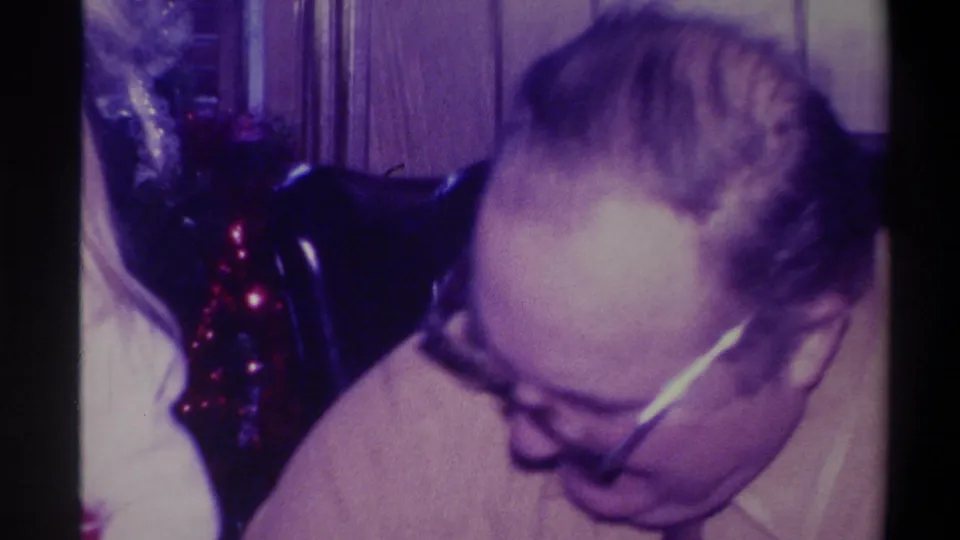 Close shot of older man with glasses looking down, Christmas tree with glowing red lights to the right, 1973 Troy New York home movie.