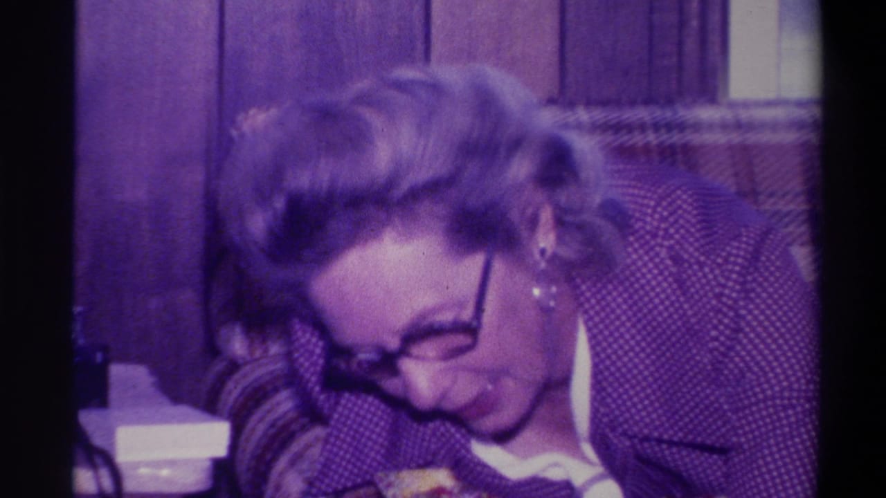 Woman with round dark-framed glasses leaning forward and looking down, wearing a dark patterned jacket, plaid couch arm in foreground, wood-paneled room.