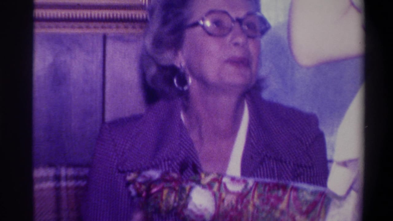 Woman in dark jacket and round glasses looking upward, holding a colorful wrapped gift; wood-paneled wall and large white rounded object at frame edge.