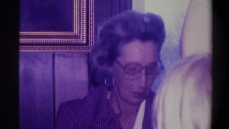 Woman in round glasses and dark jacket looking down; gold-framed picture on wood-panel wall behind her, second person's hair briefly in frame, 1973 Troy, New York.