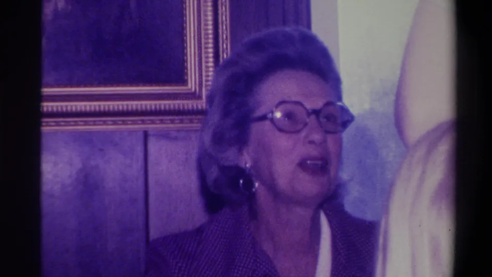 Woman in round glasses and dark jacket with mouth slightly open, speaking or reacting; gold-framed picture and wood paneling visible behind her, 1973 Troy, New York.
