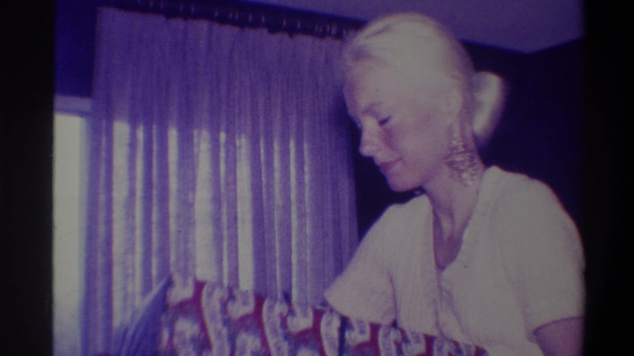 Elderly blonde woman in white short-sleeve top and ornate earrings looks downward in a domestic interior, Troy, New York, circa 1973; sheer curtains in background.