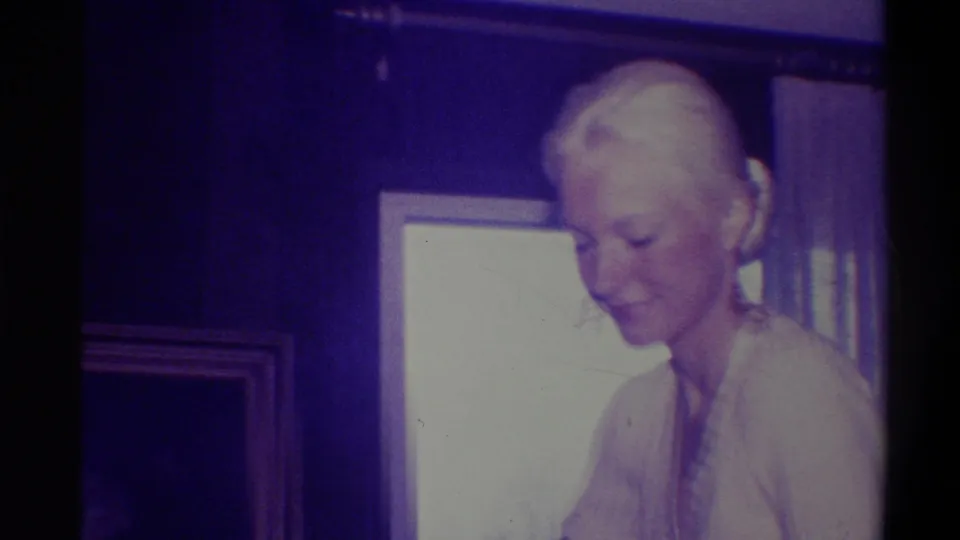 Elderly blonde woman in white top looks down in a wood-paneled room with a gold-framed picture on the wall, domestic interior circa 1973, Troy, New York.
