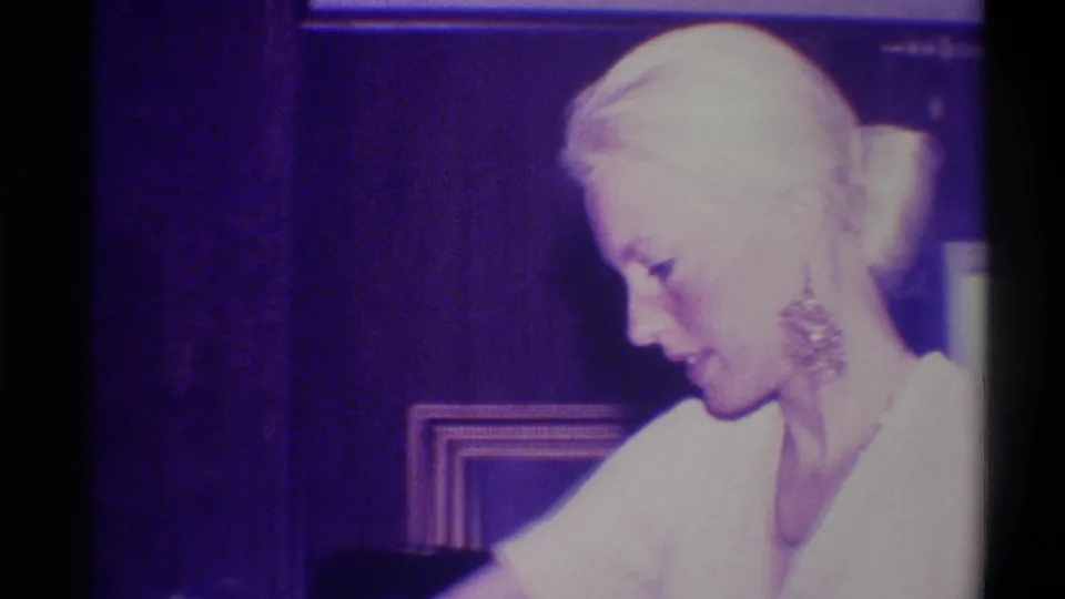 Profile close-up of an elderly blonde woman with a prominent ornate earring looking downward in a wood-paneled domestic interior, circa 1973.