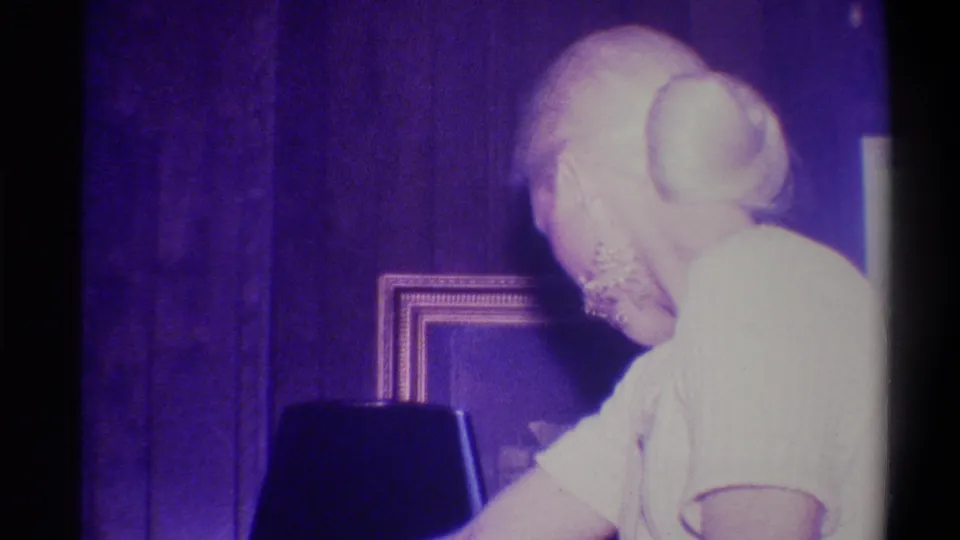 Rear-angle view of an elderly blonde woman with hair in a bun bending over a dark object in a wood-paneled domestic interior, Troy, New York, circa 1973.