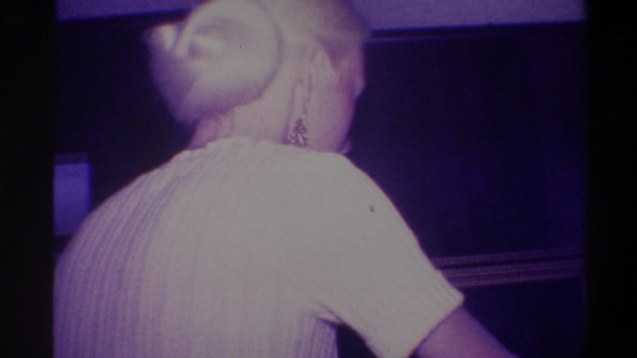 Woman with upswept blonde hair and dangling earring in a cream ribbed dress, seen from behind in a purple-lit interior, Troy, New York, circa 1973.