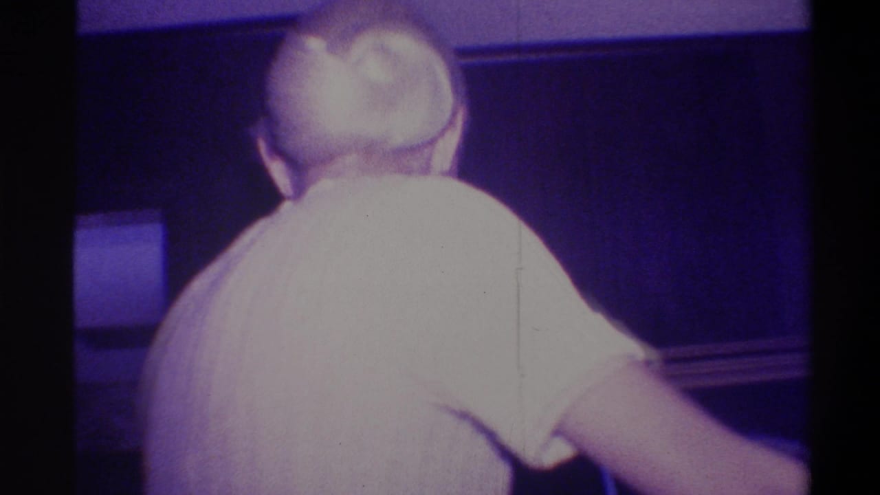 Woman with hair in a bun in a light ribbed short-sleeve dress, seen mostly from behind in a dark room with purple ambient lighting and a dark panel visible.