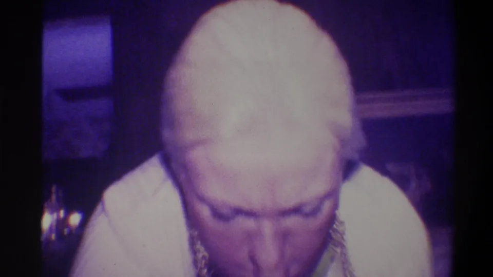 Woman bending forward in a ribbed dress, chain necklace visible, purple-lit interior with a reflective object at lower left, Troy, New York, circa 1973.
