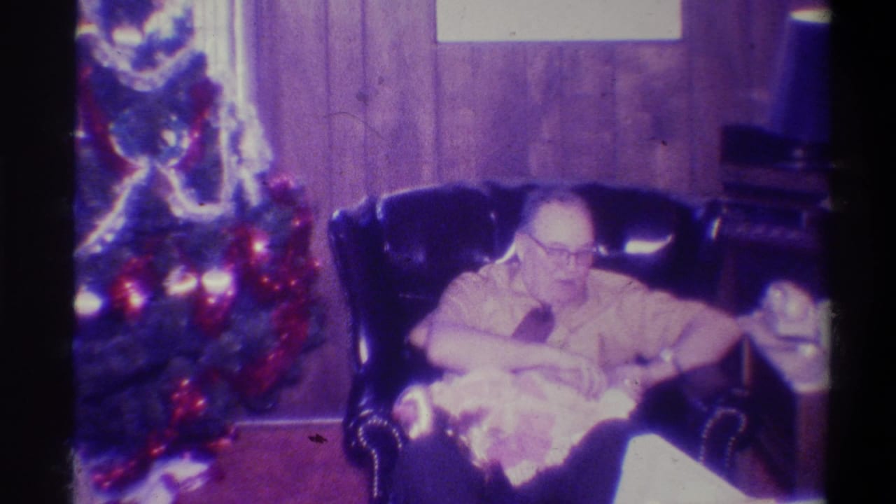 Older man in glasses seated in a dark leather armchair, Christmas tree with red ornaments to his left, wood-paneled walls, purple color cast.
