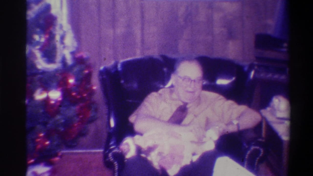 Older man with glasses in dark leather chair looking down at gift in his lap, Christmas tree with colored lights and red ornaments visible to his left.
