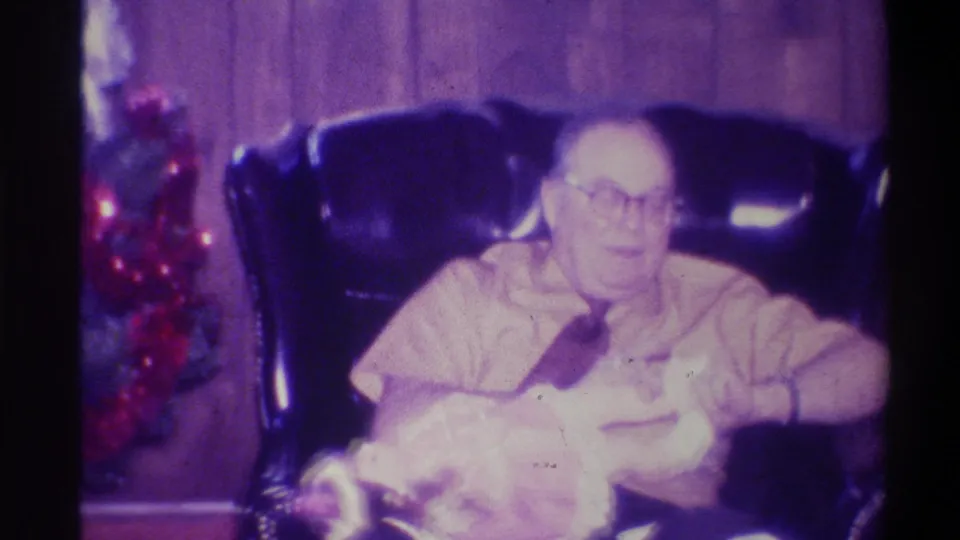 8mm home movie still, 1973 Troy New York: closer framing of older man in glasses smiling warmly in leather armchair, Christmas tree partially visible at left, film grain prominent.