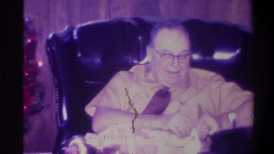 8mm home movie still, 1973 Troy New York: older man with glasses in leather armchair, emulsion scratches and film artifacts visible, Christmas tree partially at left, slight overexposure.