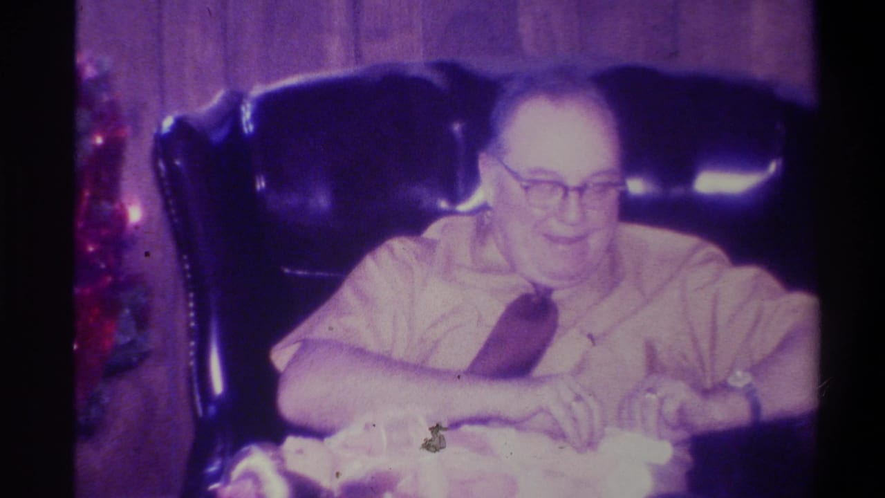 Elderly man in a leather armchair in a wood-paneled room, Troy, New York, c.1973; red holiday decorations at left frame edge; strong purple color cast from aged 8mm film.