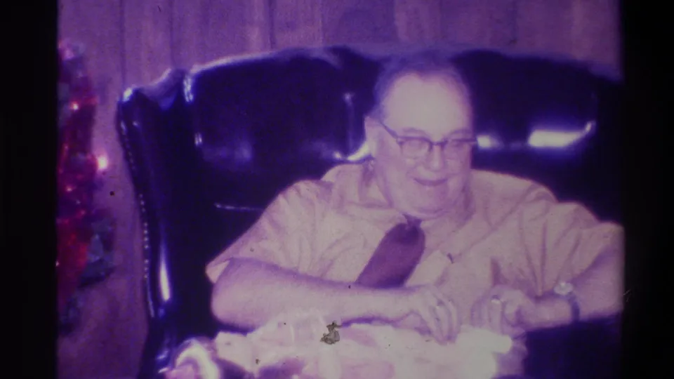 Elderly man in a leather armchair in a wood-paneled room, Troy, New York, c.1973; red holiday decorations at left frame edge; strong purple color cast from aged 8mm film.