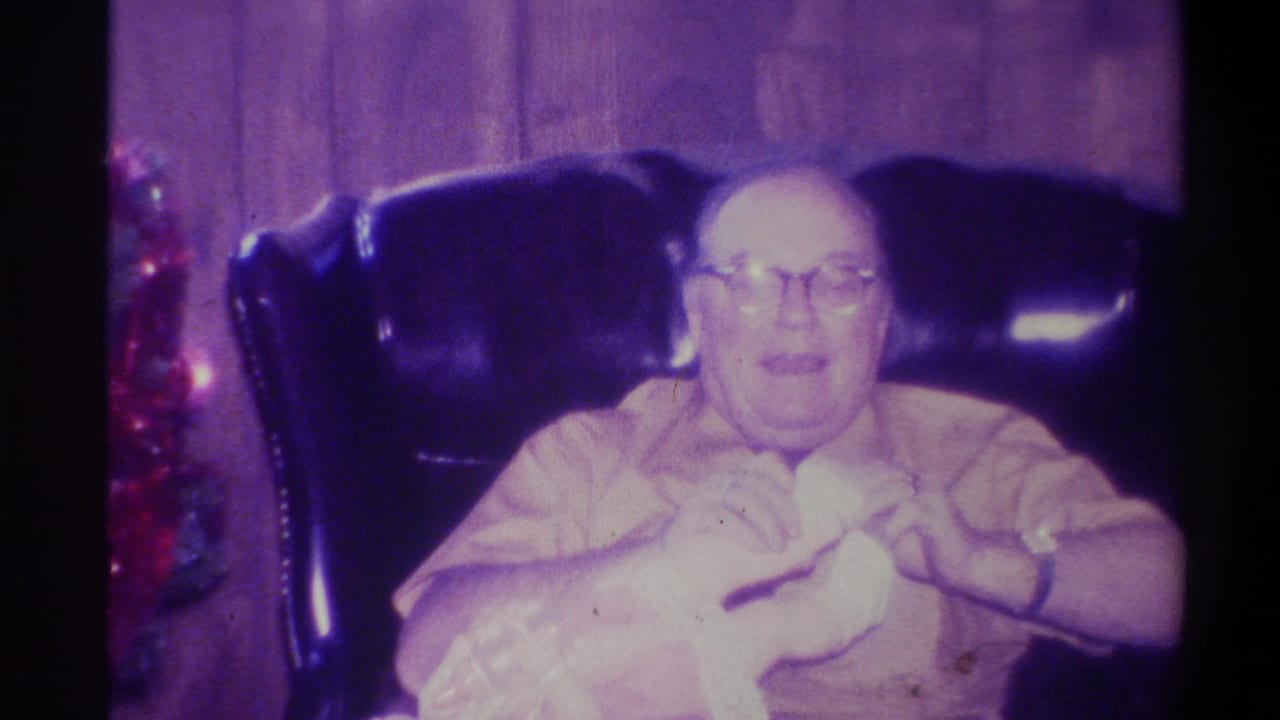 Elderly heavyset man in a leather armchair handling a white soft object in his lap; red holiday decorations visible at the right frame edge.