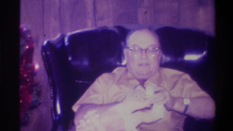 Heavyset elderly man with glasses looking toward the camera in a wood-paneled living room, Troy NY, c.1973; red decorations at frame left; 8mm purple dye-shift visible.