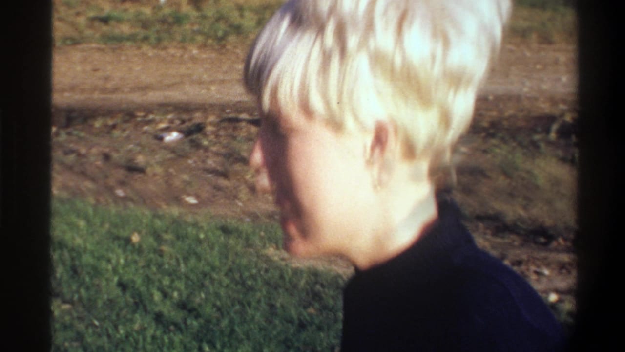 Young blonde woman seen from behind and slightly to the side, dark turtleneck, green grass and dirt road in the background.