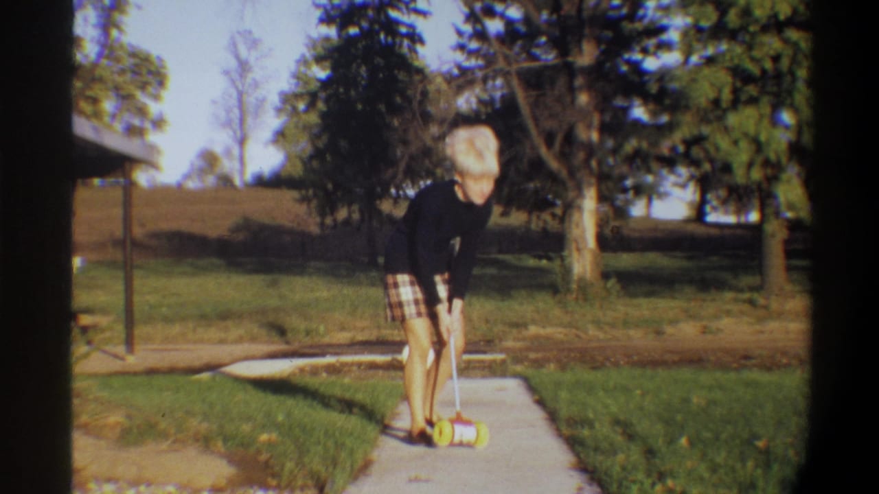 1974 home movie frame: blonde woman bending over a red-and-yellow toy on a sunlit concrete sidewalk, green lawn and trees visible behind, structure at far left.