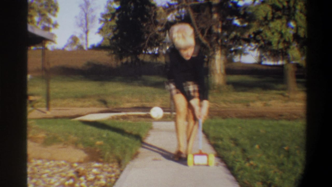 Blonde woman leans forward on concrete path guiding a small toy; gravel edging, chain-link fence, and trees visible in background.