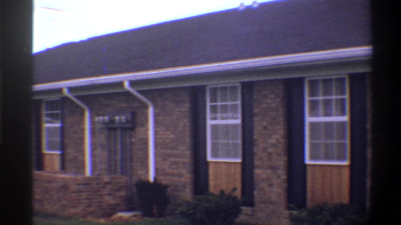8mm home movie close-up of a 1974 Gainesville, Florida brick ranch house showing dark-shuttered windows and low foundation shrubs.