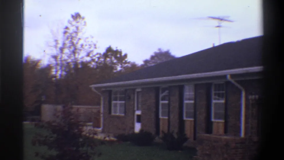 Wide 8mm shot of a 1974 Gainesville, Florida brick ranch house; TV antenna silhouetted against overcast sky with mature trees behind.