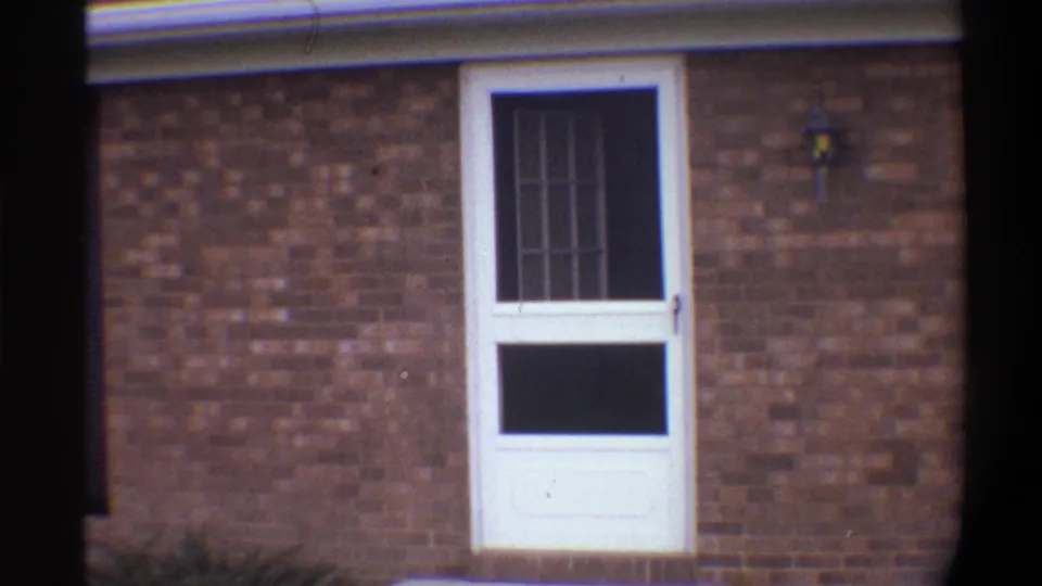 Close-up 8mm frame of a white screen door with security grid grille in a brick ranch house facade, circa 1974 Gainesville, Florida.