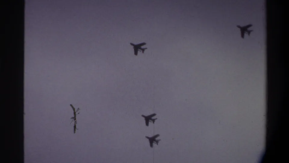 Jet formation continuing across overcast sky on 8mm film, 1974, Gainesville, Florida; a bare tree tip intrudes at frame edge.
