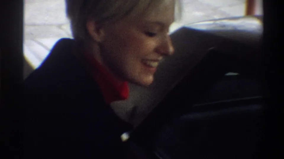 Close-up of a smiling woman with short blonde hair in a dark coat and red turtleneck, filmed on 8mm in Gainesville, Florida, circa 1974.