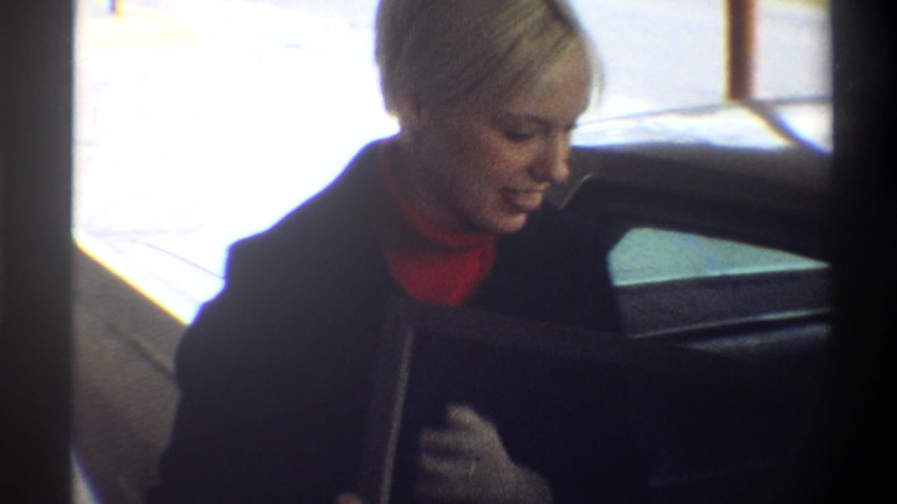 Woman in dark coat and red turtleneck at open car door, overexposed outdoor background, 8mm home movie.