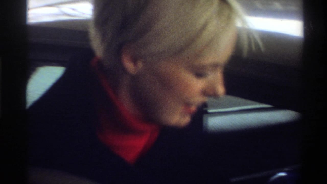 Close-up of woman with short blonde hair in dark coat and red turtleneck looking downward, 8mm home movie, Gainesville, Florida, circa 1974.