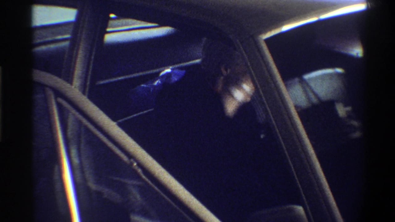 Looking through an open car door into a dark interior with chrome door trim; a figure is partially visible inside, filmed on 8mm circa 1974.