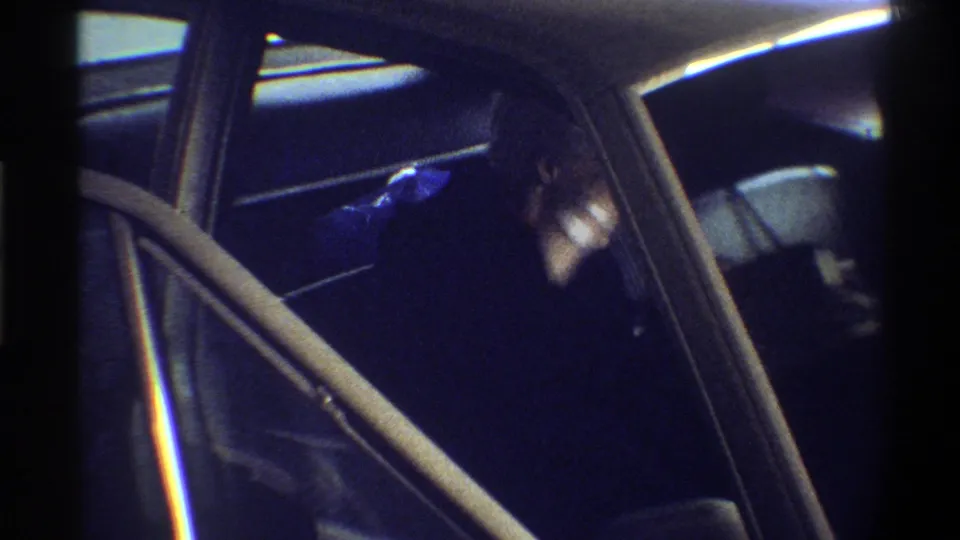 Looking through an open car door into a dark interior with chrome door trim; a figure is partially visible inside, filmed on 8mm circa 1974.