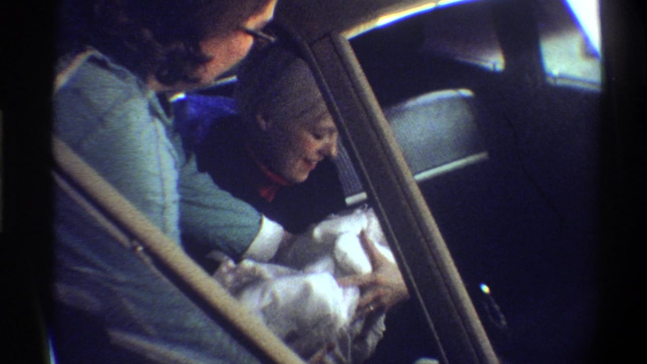 Woman cradling a newborn wrapped in white blankets in a car backseat, 8mm home movie, Gainesville, Florida, circa 1974.
