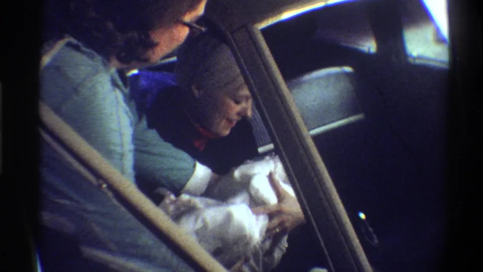 Woman cradling a newborn wrapped in white blankets in a car backseat, 8mm home movie, Gainesville, Florida, circa 1974.