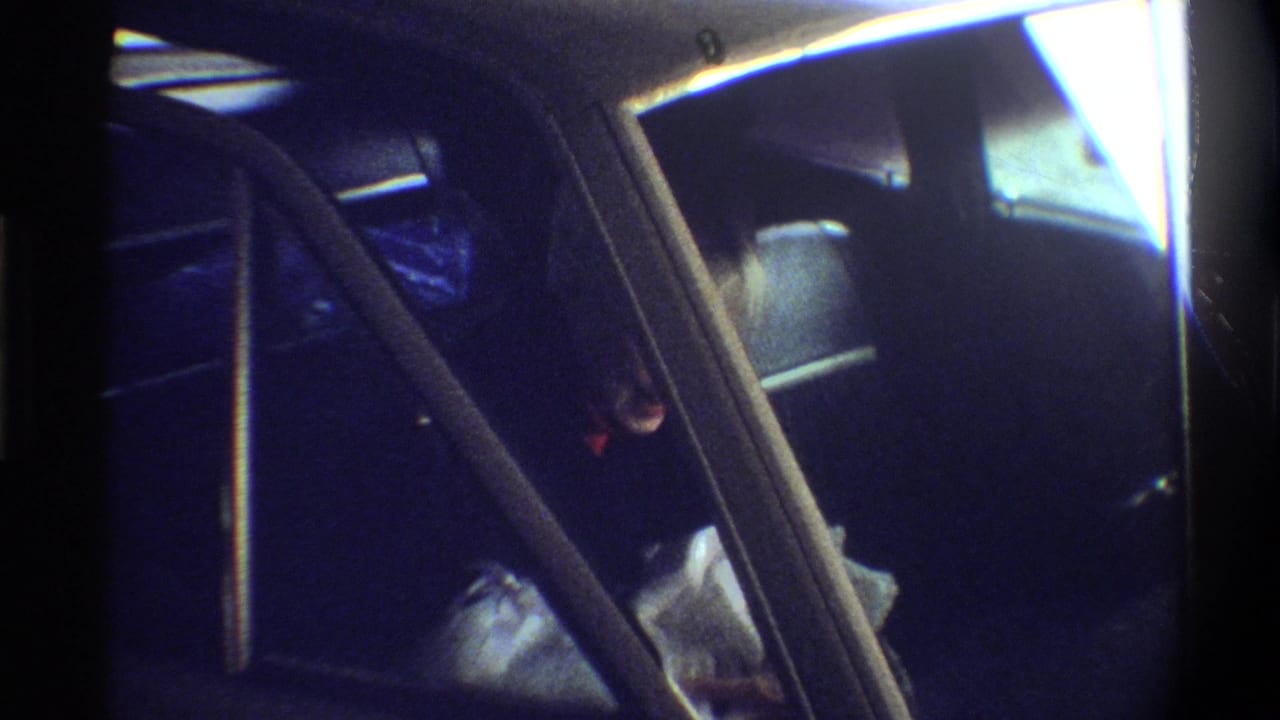 Car backseat showing newborn wrapped in white blankets alongside a blue bag; woman partially visible at left edge; 8mm home movie, circa 1974.