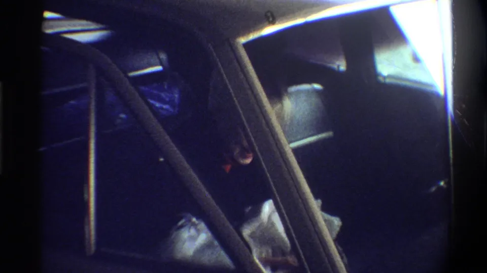 Car backseat showing newborn wrapped in white blankets alongside a blue bag; woman partially visible at left edge; 8mm home movie, circa 1974.
