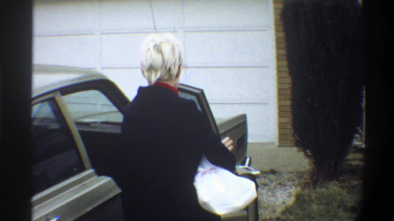 Woman with short blonde hair and red collar in dark jacket standing at open car door; white-panelled wall and brick exterior behind; 8mm home movie, Gainesville, Florida, circa 1974.