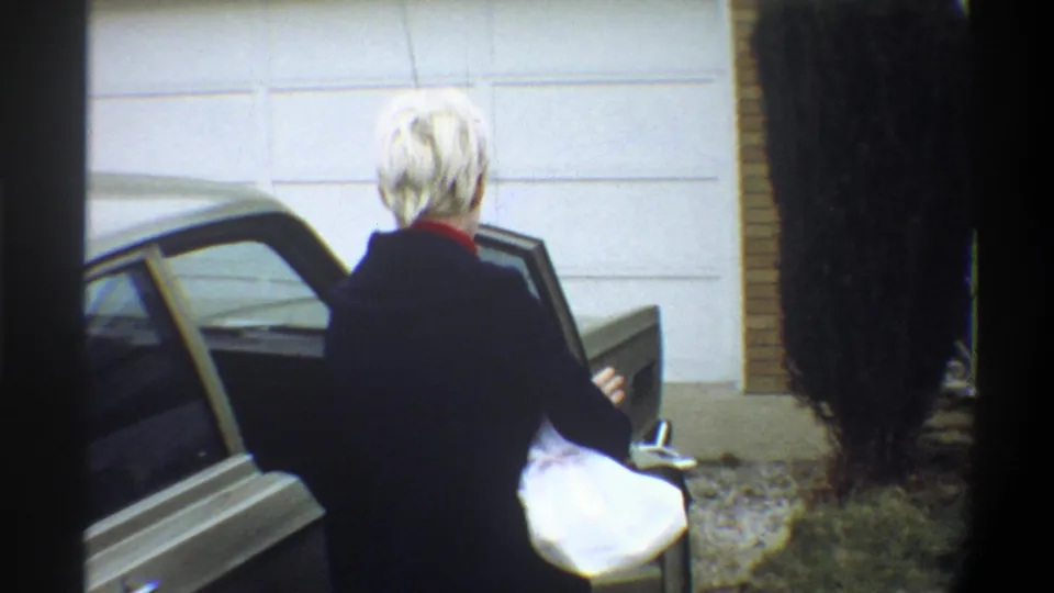 Woman with short blonde hair and red collar in dark jacket standing at open car door; white-panelled wall and brick exterior behind; 8mm home movie, Gainesville, Florida, circa 1974.