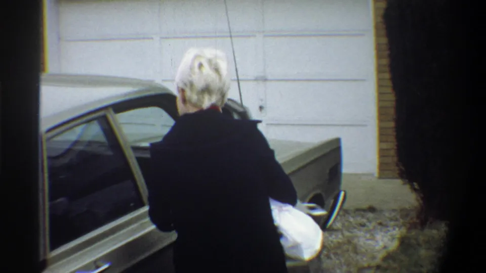 Woman in dark coat exits a 1970s olive-gold sedan holding what appears to be a bundled infant, suburban Gainesville FL, circa 1974.