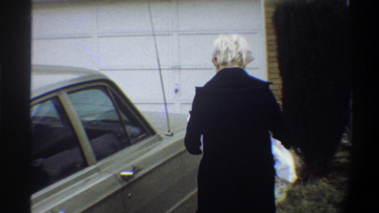 Woman in dark coat carries a white-wrapped bundle away from a parked 1970s sedan toward a white suburban garage door, Gainesville FL, circa 1974.