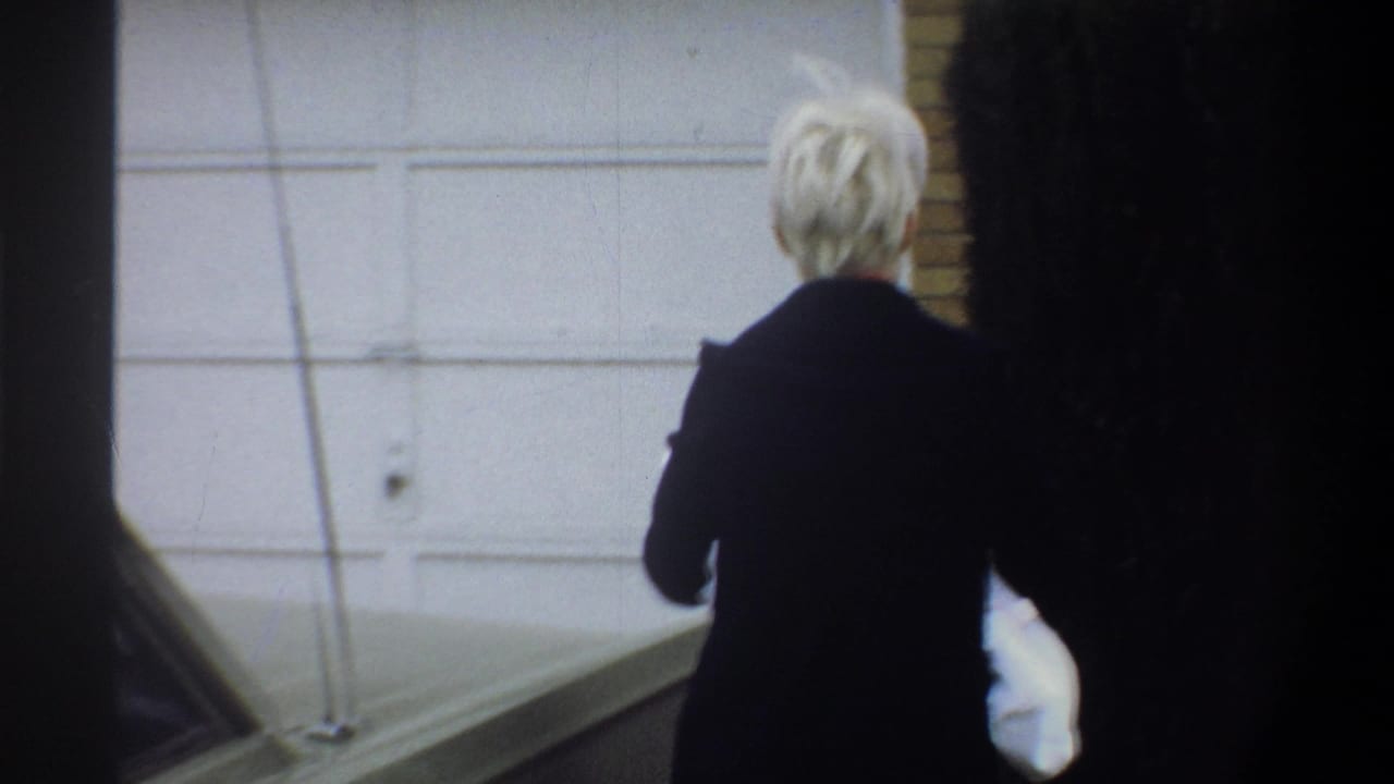 Woman in dark coat approaches a white suburban garage door carrying a white-wrapped bundle, 8mm home movie, Gainesville FL, circa 1974.