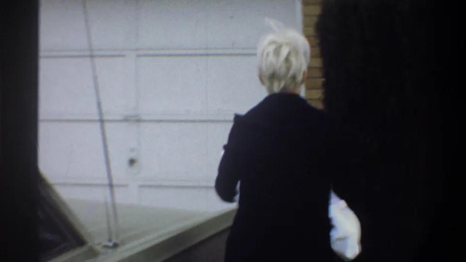 Woman in dark coat approaches a white suburban garage door carrying a white-wrapped bundle, 8mm home movie, Gainesville FL, circa 1974.