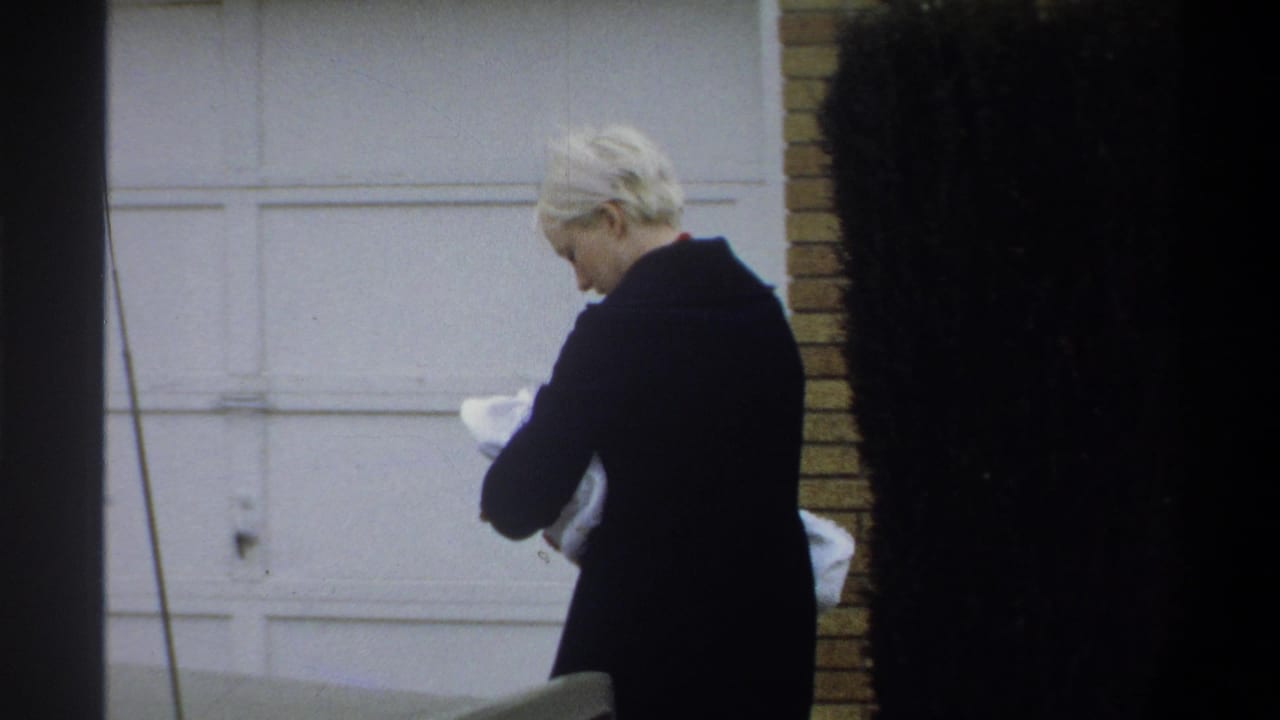 Woman in dark coat seen in partial profile, looking down at a white-wrapped bundle, brick wall and white garage door visible, Gainesville FL, circa 1974.
