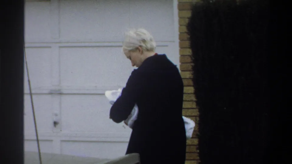 Woman in dark coat seen in partial profile, looking down at a white-wrapped bundle, brick wall and white garage door visible, Gainesville FL, circa 1974.
