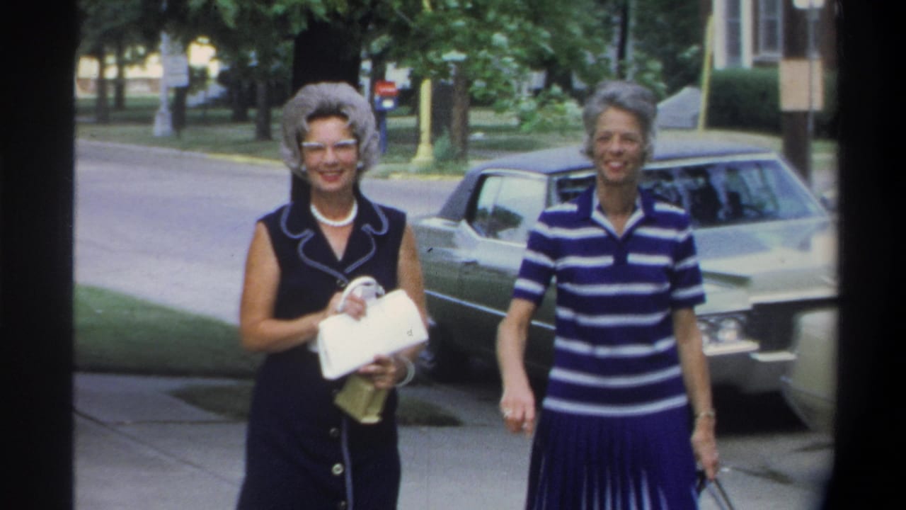 Two well-dressed older women pose on a suburban sidewalk in footage described as Gainesville, Florida, 1974; a large cream sedan and red car are visible in the background.