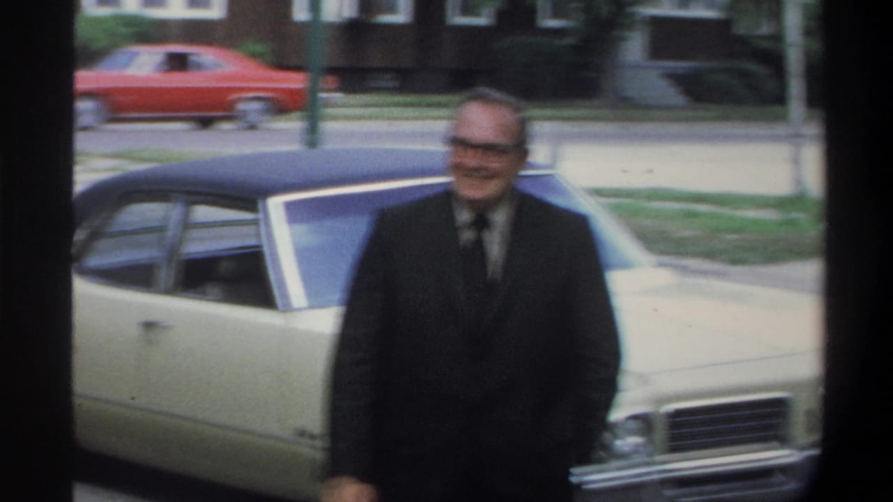 An older man in glasses and a dark suit smiles beside a cream American sedan in footage described as Gainesville, Florida, circa 1974; a red car and suburban trees are visible behind him.