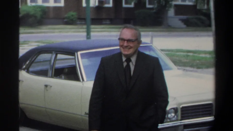 An older man in glasses and a dark suit smiles warmly beside the front of a cream American sedan; suburban trees and a concrete sidewalk are visible, consistent with early-1970s home-movie footage.
