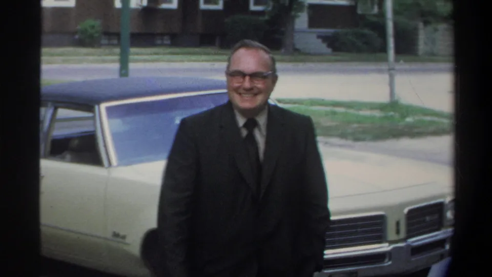 An older man in glasses and a dark suit smiles at the camera beside a cream sedan; this frame closely mirrors the preceding one, consistent with continuous handheld 8mm shooting in Gainesville, Florida, circa 1974.