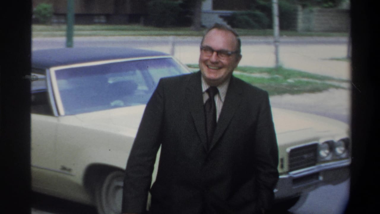 Man in dark suit, dark tie, and wire-frame glasses smiling beside a large cream American sedan; residential street with greenery behind him, Gainesville Florida area, circa 1974.